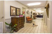 A clean, well-lit hallway in a nursing and rehab center with tiled floors, a wooden chest of drawers with decorative items and framed artwork on the wall above it, a potted plant on the floor, and a couple of chairs along the wall. The hallway leads to a reception or nurse's station area in the background.