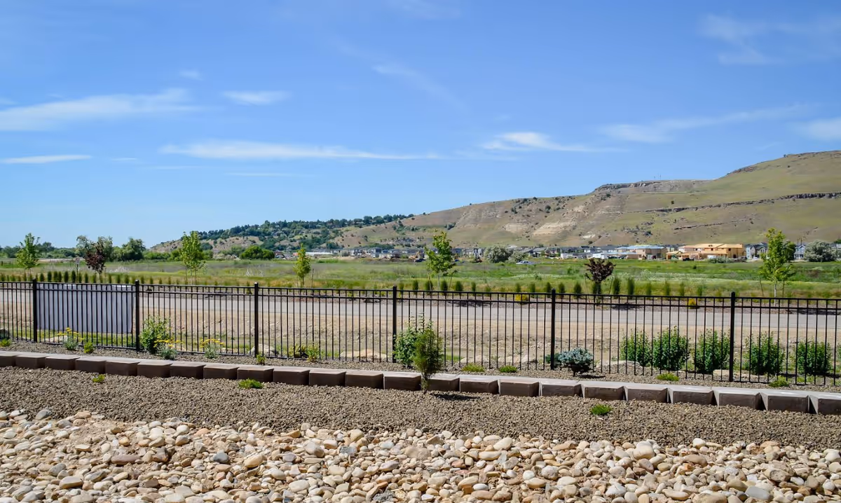 View of a fenced yard with gravel and rocks overlooking grassy fields and distant hills under a blue sky.