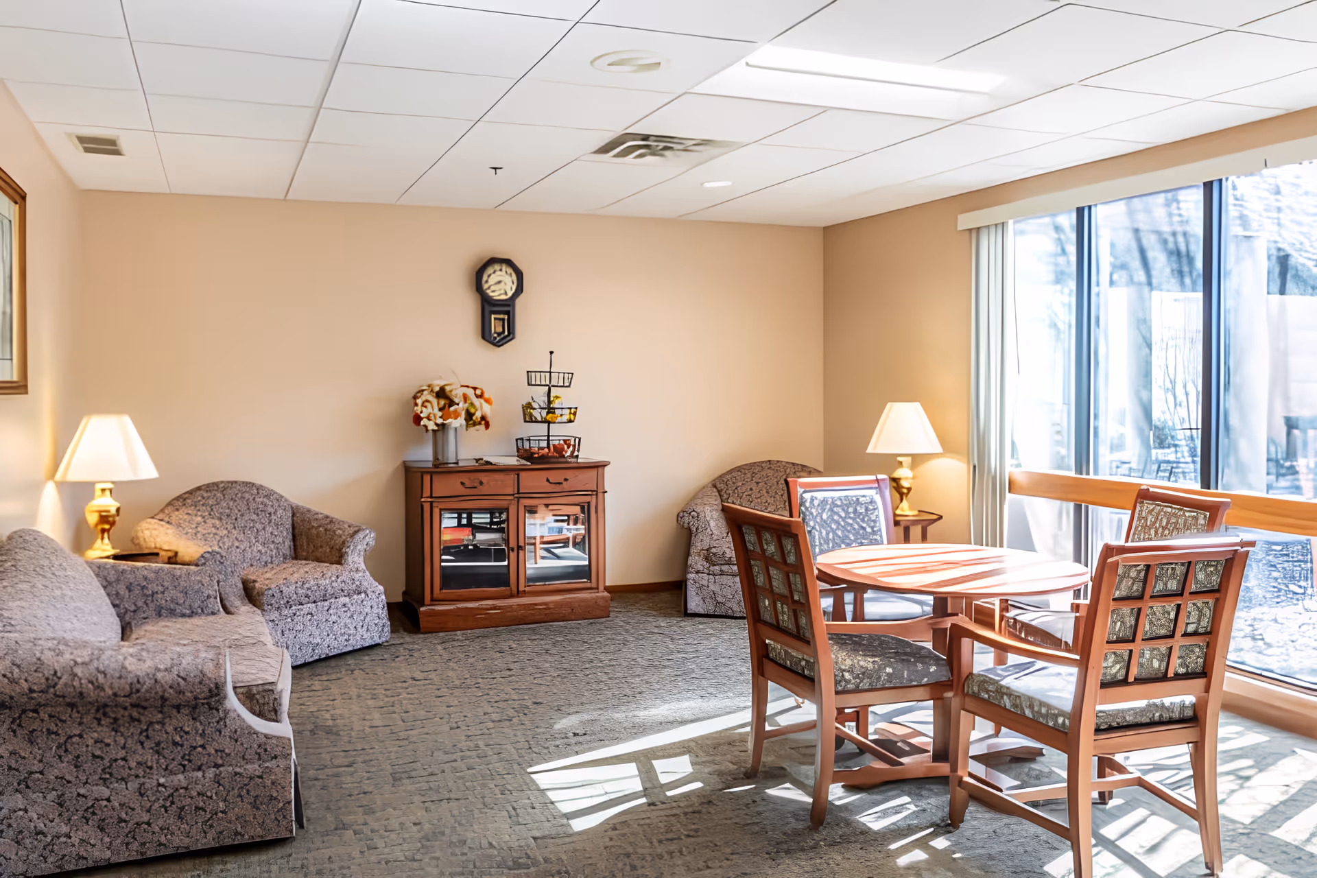 Sunlit communal lounge with sofas and armchairs, a round table with four chairs by large windows, lamps, and a small cabinet.