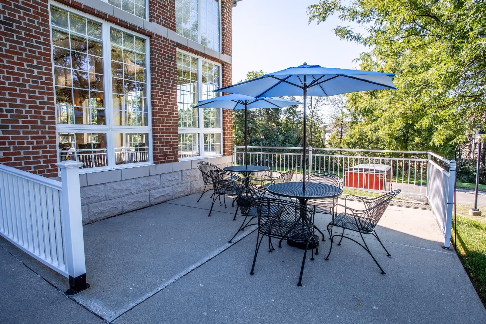 Outdoor patio area with two round metal tables and several metal chairs, each table shaded by a blue umbrella. The patio is adjacent to a brick building with large windows, surrounded by a white railing and greenery in the background.