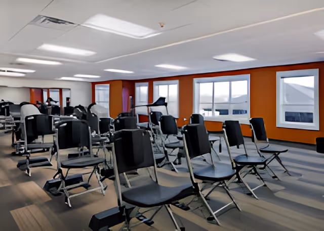 A spacious room with multiple rows of black chairs arranged facing forward, large windows letting in natural light, and orange accent walls. The ceiling has recessed lighting, and there is a mirrored wall on one side.