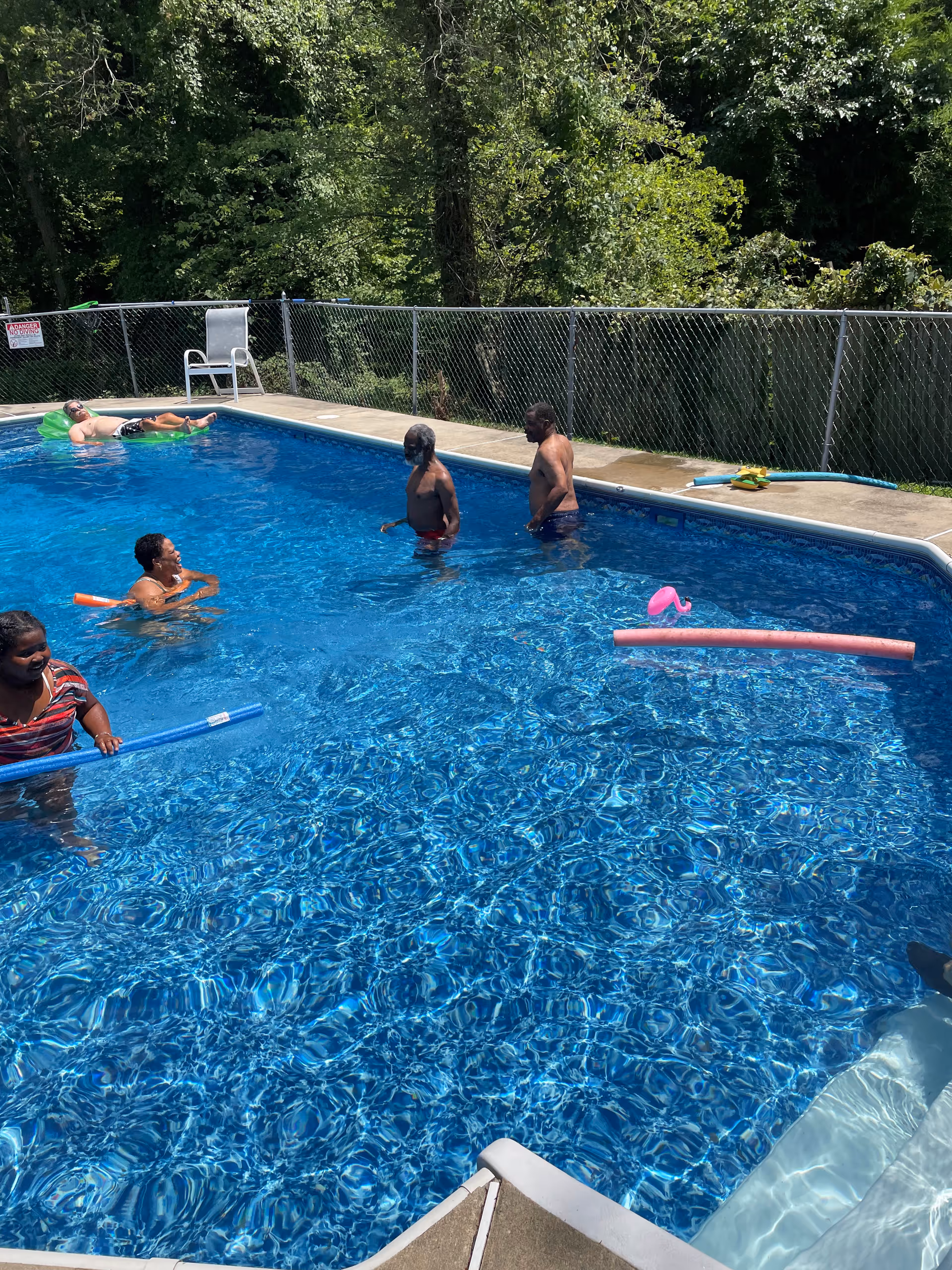 Several people enjoying a sunny day in an outdoor swimming pool surrounded by a chain-link fence and trees in the background. Some individuals are standing or swimming in the pool, while one person is floating on a green inflatable. Pool noodles and a pink flamingo float are also visible in the water.