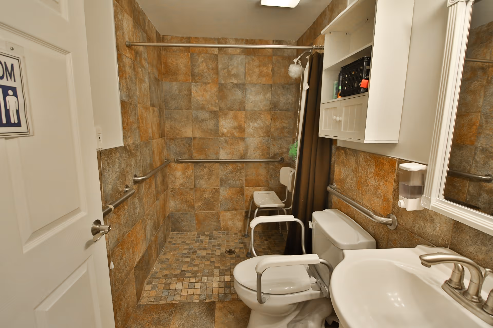 A bathroom with brown and beige tiled walls and floor. It features a walk-in shower with a shower chair and grab bars, a toilet with arm supports, a white sink with a silver faucet, a wall-mounted soap dispenser, and a white cabinet with shelves above the toilet. The door to the bathroom is open, showing a restroom sign.