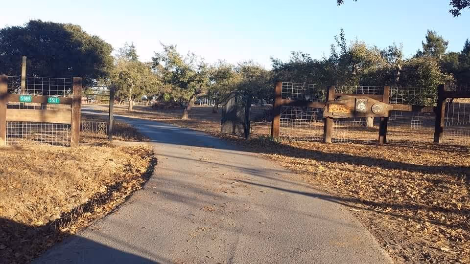 A paved driveway leading through a wooden gate with wire fencing on both sides, surrounded by dry grass and trees under a clear sky.