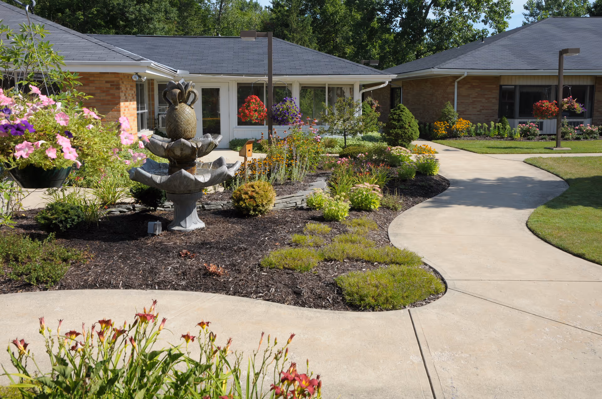 Sunlit courtyard with a tiered pineapple fountain, winding concrete paths, and flowerbeds in front of a single-story brick senior living building.