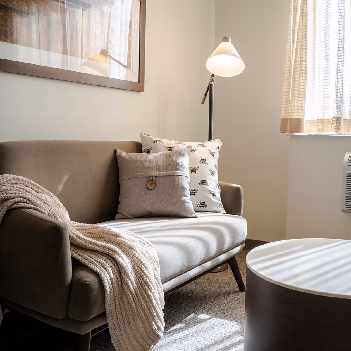 A cozy living area with a beige loveseat draped with a knitted throw, decorative pillows, a floor lamp, and a round coffee table by a sunlit window.