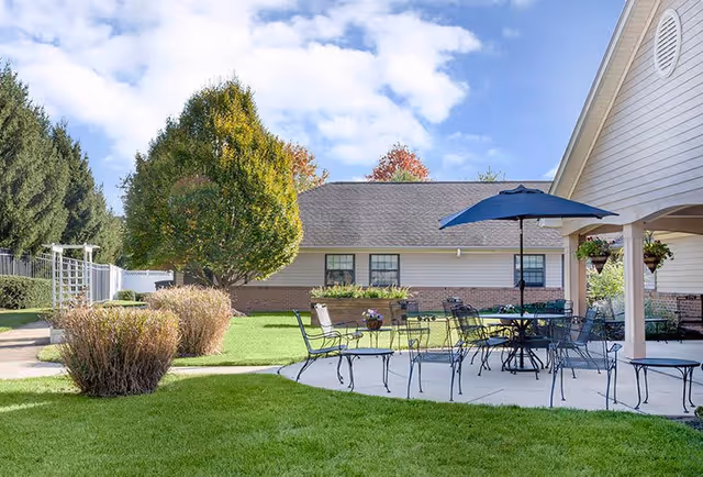 Outdoor patio area at a senior living facility with metal tables and chairs, one table shaded by a large blue umbrella. The patio is surrounded by green grass, bushes, and trees with a building in the background under a partly cloudy sky.