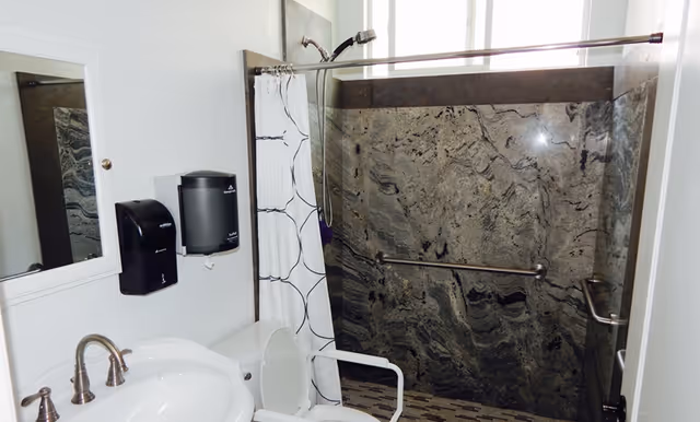 A senior-friendly bathroom featuring a white sink with silver faucets, a toilet with a white safety frame, a shower area with a patterned curtain and dark marble walls, a showerhead, and grab bars for support. There are also wall-mounted soap and paper towel dispensers next to a mirror.