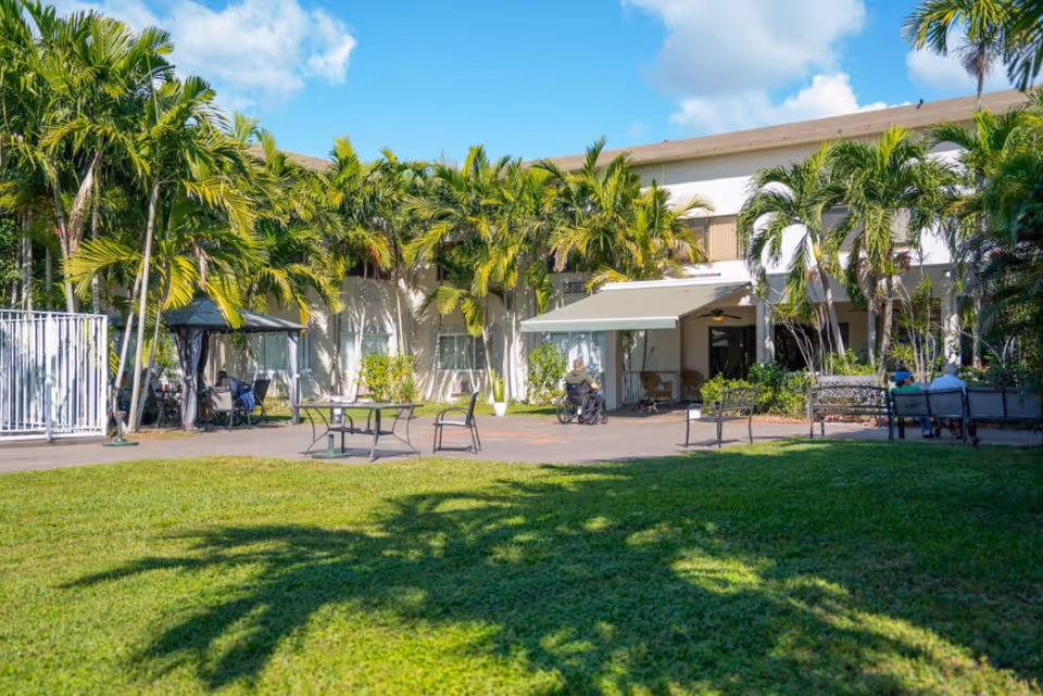 Outdoor courtyard area of a senior living facility with green grass, palm trees, patio tables and chairs, and a few seniors sitting on benches and in a wheelchair under a covered area attached to a two-story building.