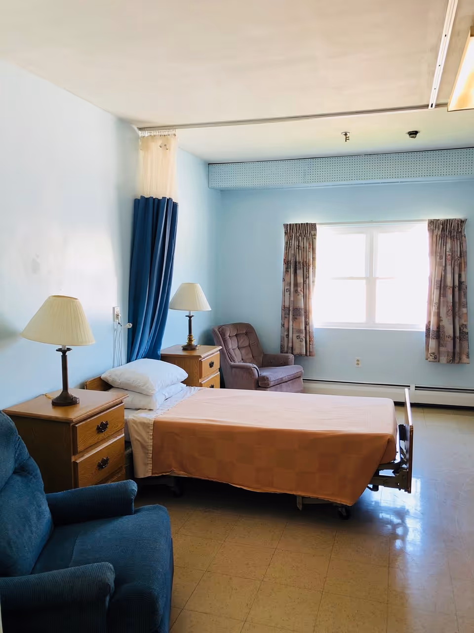 A simple, clean bedroom in a senior living facility with a single hospital-style bed covered with a peach blanket, two wooden nightstands each with a lamp, a blue armchair, a brown cushioned chair near a window with floral curtains, and light blue walls.