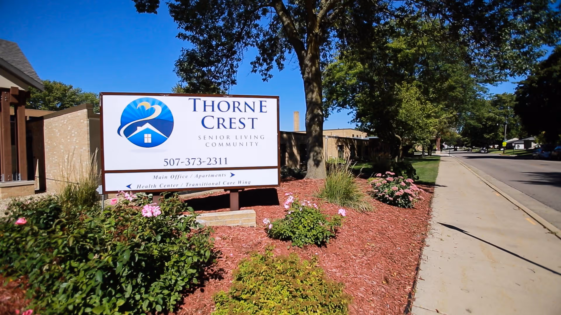 Sign for Thorne Crest Senior Living Community in a landscaped front bed beside a sidewalk and street.