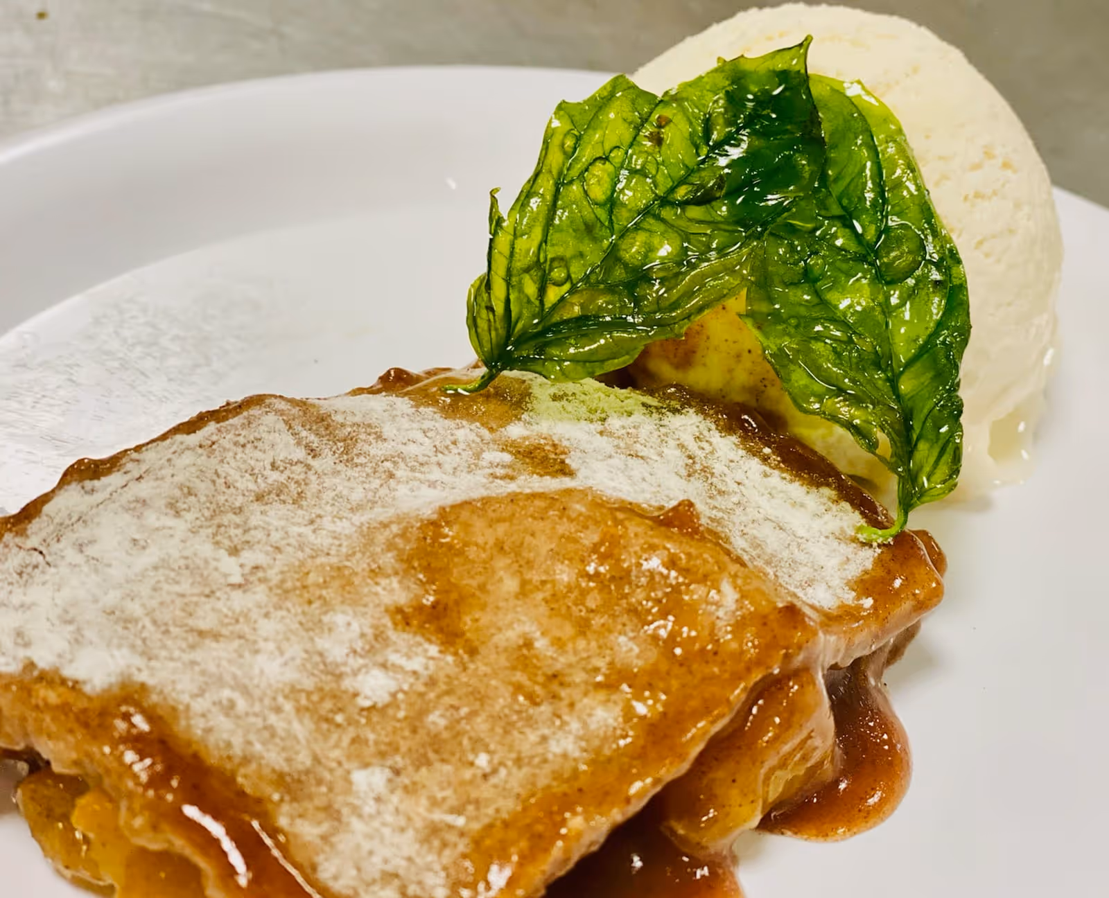A close-up of a dessert plate featuring a slice of apple pie dusted with powdered sugar, topped with a glazed green leaf, and served with a scoop of vanilla ice cream.