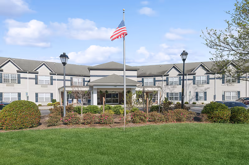 Front exterior view of a two-story senior living facility building with multiple windows, a covered entrance, an American flag on a flagpole, two street lamps, and landscaped bushes and grass in the foreground under a partly cloudy sky.