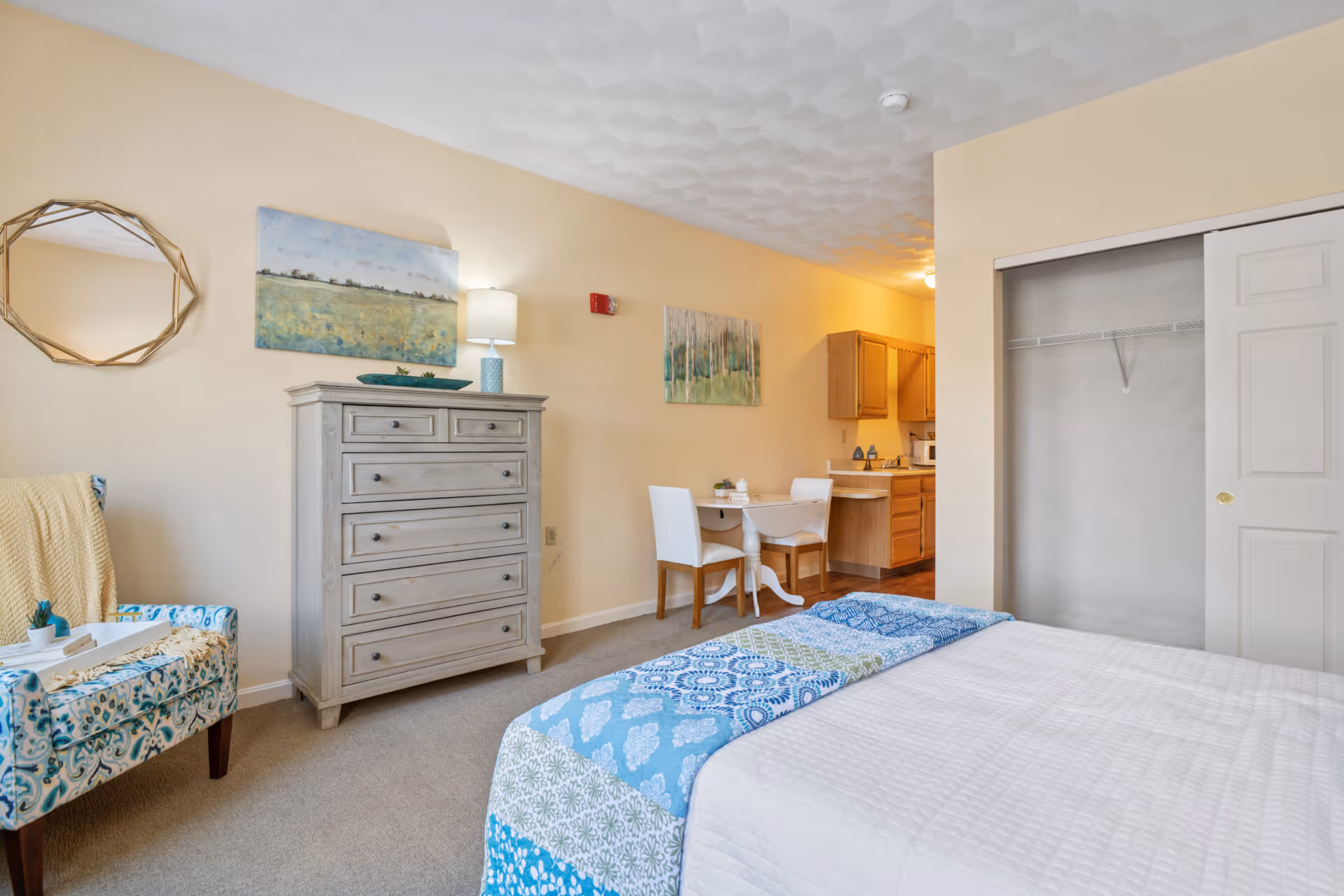 A cozy bedroom with a bed covered in a white quilt and a blue patterned throw. To the left is a patterned armchair with a yellow blanket draped over it and a small tray on the armrest. A gray chest of drawers stands against the wall with a lamp and decorative items on top. Above the chest are two paintings and a round mirror. In the background, there is a small dining table with two white chairs and a kitchenette with wooden cabinets. An open closet with sliding doors is visible on the right side.