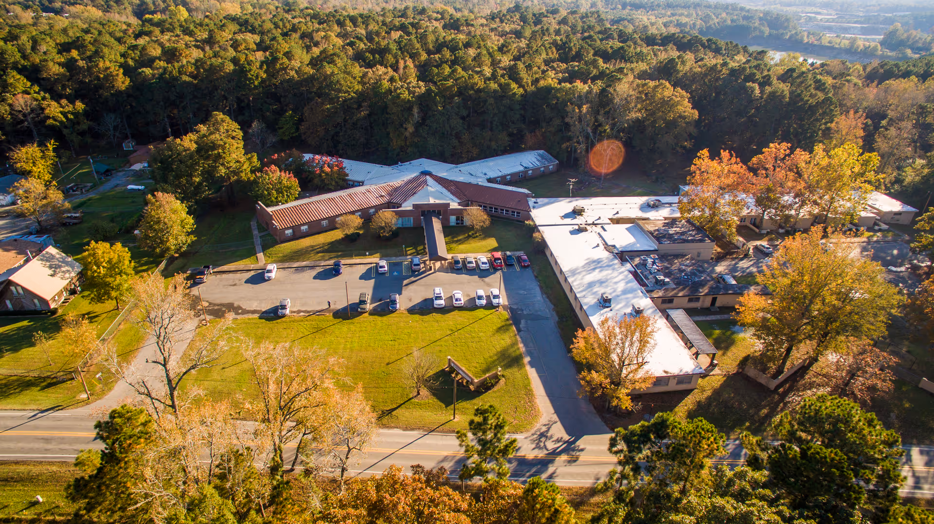 Aerial view of The Blossoms at West Dixon Assisted Living, Nursing Center & Rehab facility surrounded by trees with autumn foliage. The building complex includes multiple connected structures with parking spaces in front and a road running along the front of the property.