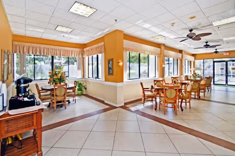 Bright communal dining area with round tables and chairs, large windows, ceiling fans, and a coffee station.