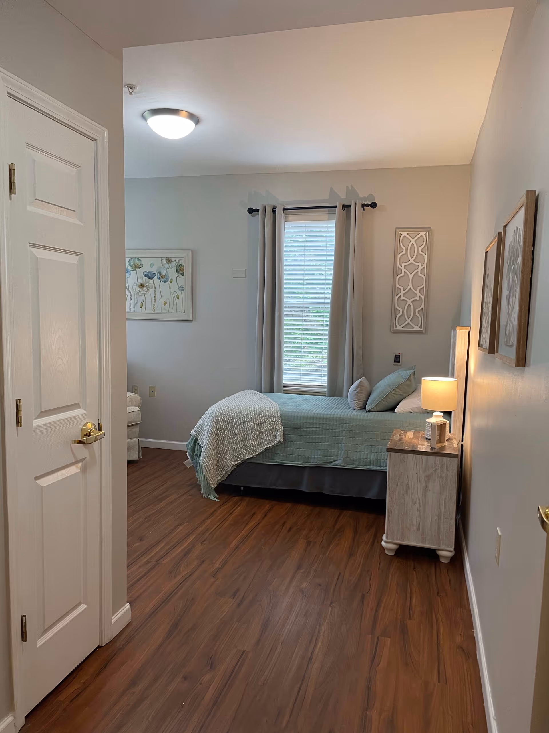 A cozy bedroom with a single bed covered in a light green quilt and a textured throw blanket. There are two pillows on the bed. Next to the bed is a wooden nightstand with a lamp and a small decorative item. The room has wooden flooring, a window with closed blinds and light gray curtains, and light-colored walls adorned with framed artwork. A white door is partially open on the left side of the image.