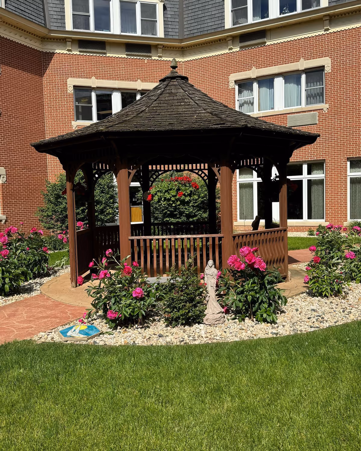 A wooden gazebo with a shingled roof situated in a garden area with green grass, pink flowers, and a small statue. The gazebo is surrounded by a circular stone and flower bed, with a red brick building in the background featuring multiple windows.