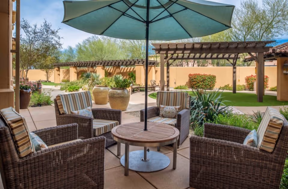 Outdoor patio area with four wicker chairs with striped cushions arranged around a round wooden table with a large green umbrella. The patio is surrounded by plants, large decorative pots, and a pergola structure in the background with a beige wall and trees beyond.