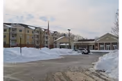 Snow-covered driveway and piled snow in front of a multi-story beige senior living building with an entrance canopy and a parked car.