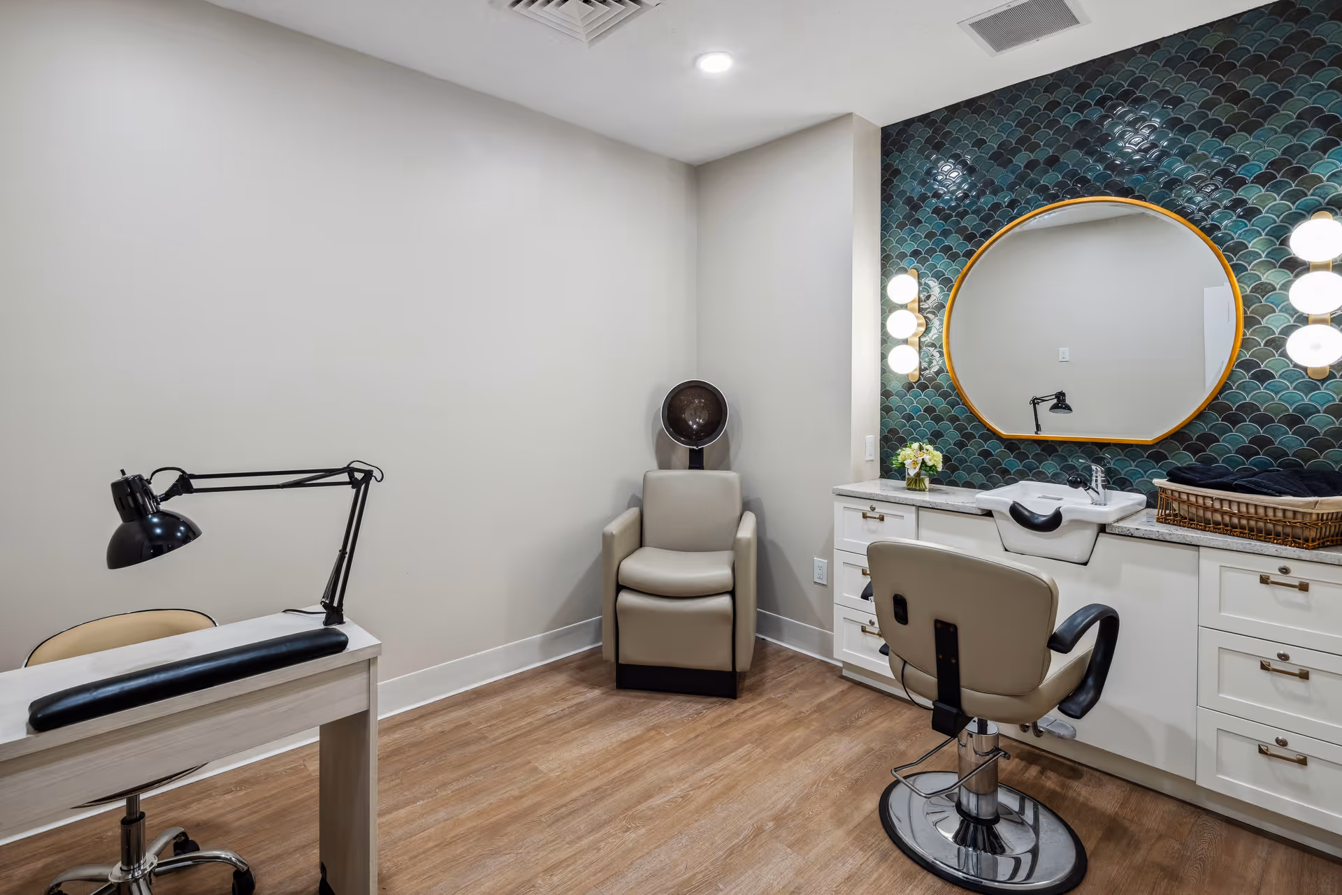 Salon-style interior with a styling chair, wash sink under a round mirror, tiled accent wall, and a manicure table.
