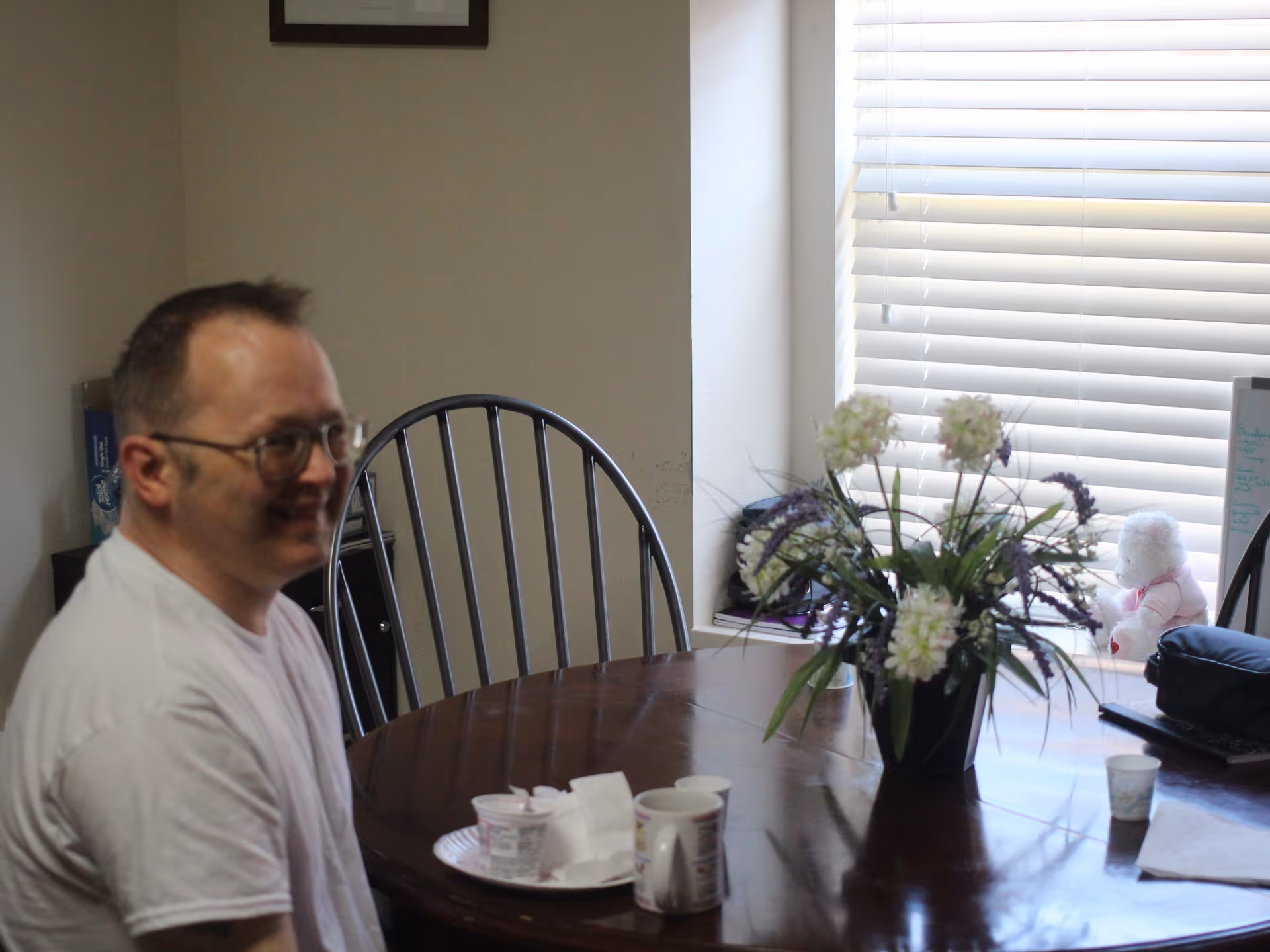 A man wearing glasses and a white shirt sitting at a wooden dining table with a vase of flowers, cups, and a plate. The room has a window with blinds and a teddy bear on the windowsill.