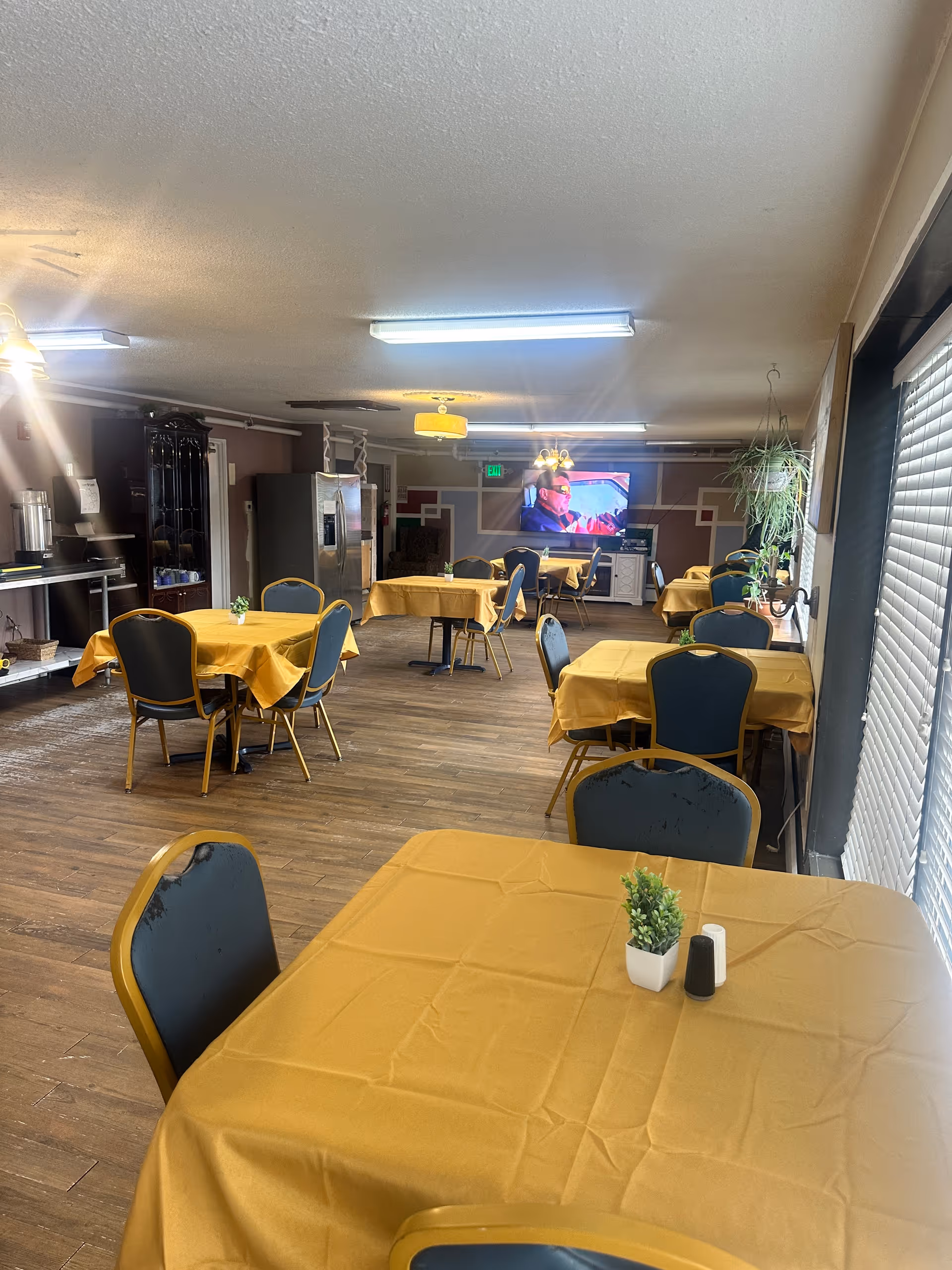 Interior view of a dining room in an assisted living facility with several tables covered in yellow tablecloths and blue chairs around them. There is a small plant and salt and pepper shakers on the closest table. The room has wooden flooring, a television mounted on the far wall, and large windows with blinds on the right side. Various light fixtures hang from the ceiling, and there are some plants and furniture along the walls.