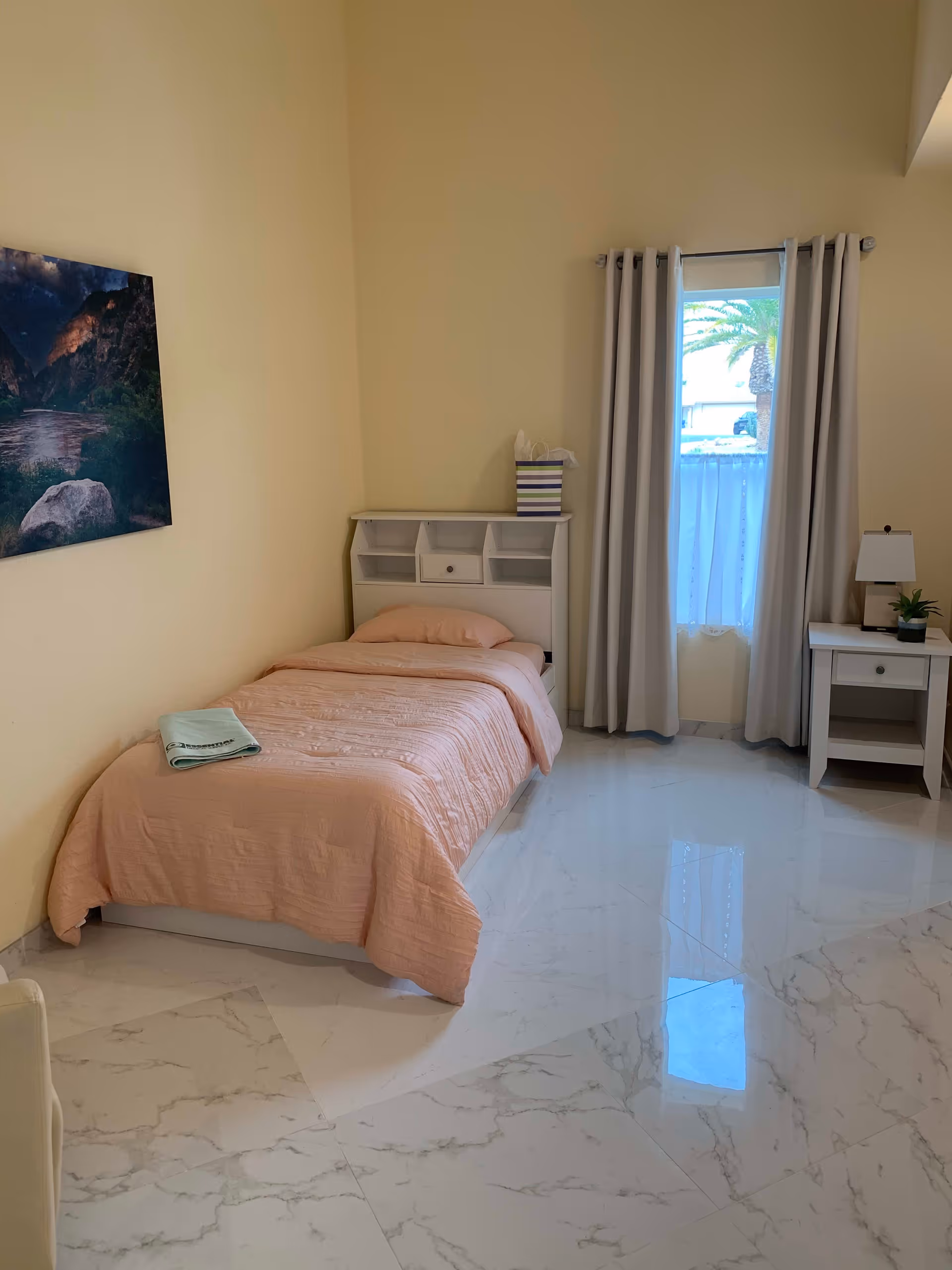 A simple bedroom with a single bed covered in a peach-colored quilt, a white headboard with shelves, a small white nightstand with a lamp and a small plant, a window with beige curtains, and a landscape painting on the wall. The floor is white marble tile.