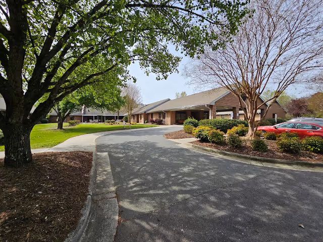 View of the front exterior of Alamance Health Care Center showing a curved driveway leading to the entrance. The building is a single-story structure with a brick facade and a covered entrance. There are trees with green and bare branches, landscaped bushes, and a red car parked near the entrance under a clear sky.