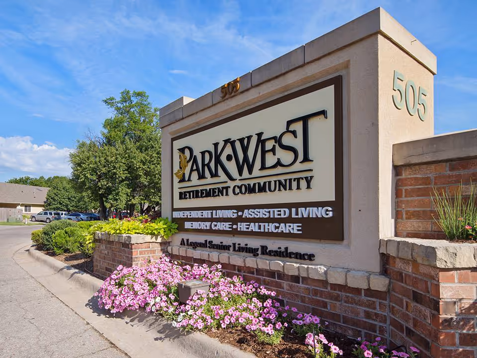 Entrance sign for Park West Retirement Community with text indicating Independent Living, Assisted Living, Memory Care, and Healthcare services. The sign is mounted on a brick and stone structure with pink flowers planted at the base. Trees, parked cars, and a clear blue sky are visible in the background.
