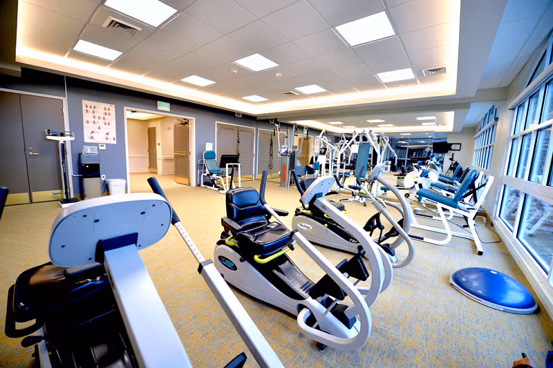 Bright and spacious fitness room with various exercise equipment including recumbent bikes, elliptical machines, and weight machines. Large windows on the right side allow natural light to fill the room. The ceiling has recessed lighting and the floor is carpeted. There are doors and a hallway visible at the far end of the room.