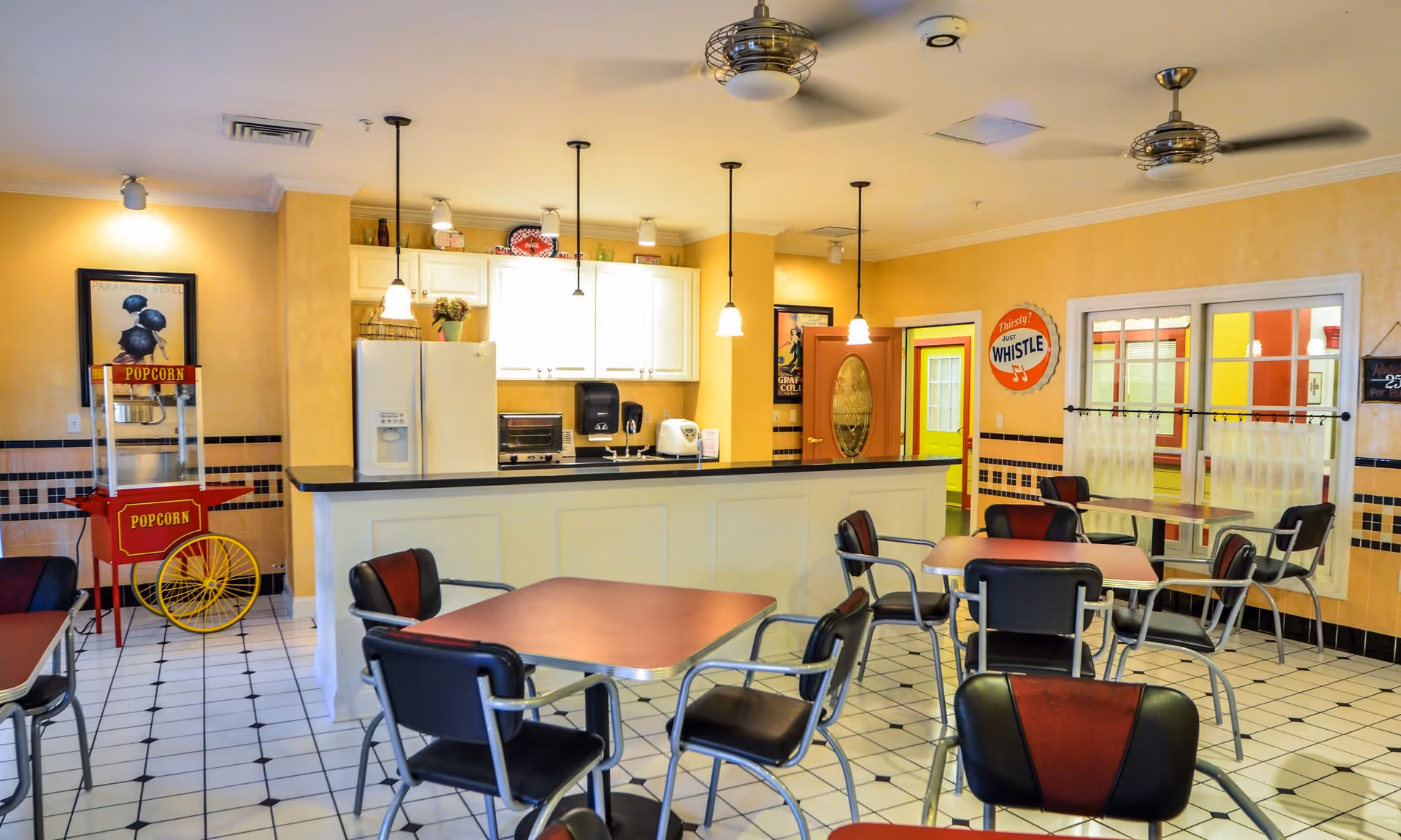 A bright dining area with several tables and chairs, a popcorn machine on the left, and a kitchen counter with white cabinets and appliances in the background. The room has yellow walls, black and white tiled floor, and ceiling fans with lights.