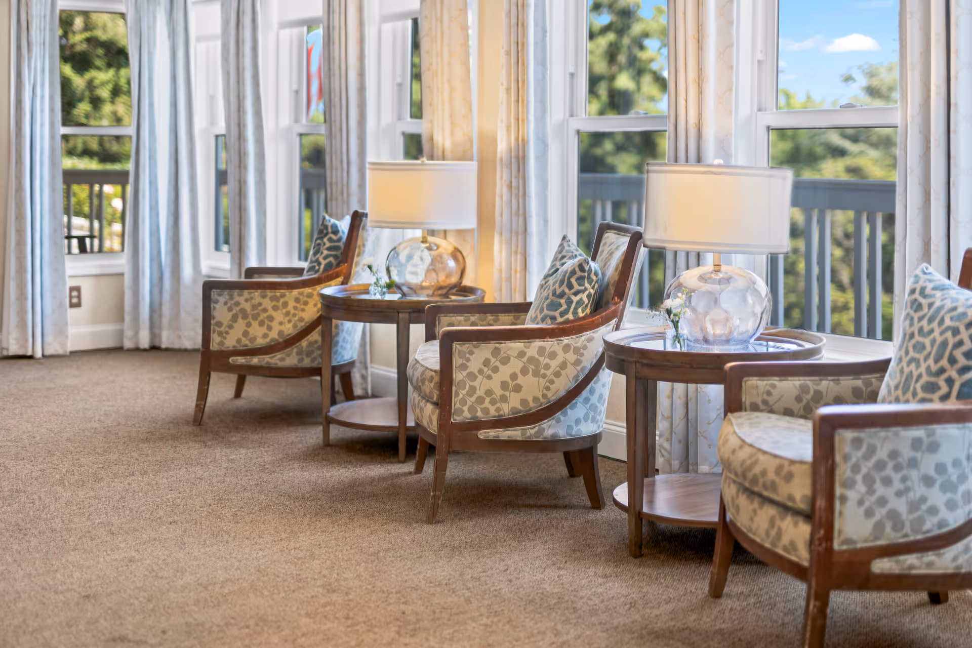 A cozy seating area with three upholstered armchairs featuring patterned cushions, each paired with a round wooden side table holding a glass lamp and small flower vase. Large windows with light curtains provide a view of greenery outside.