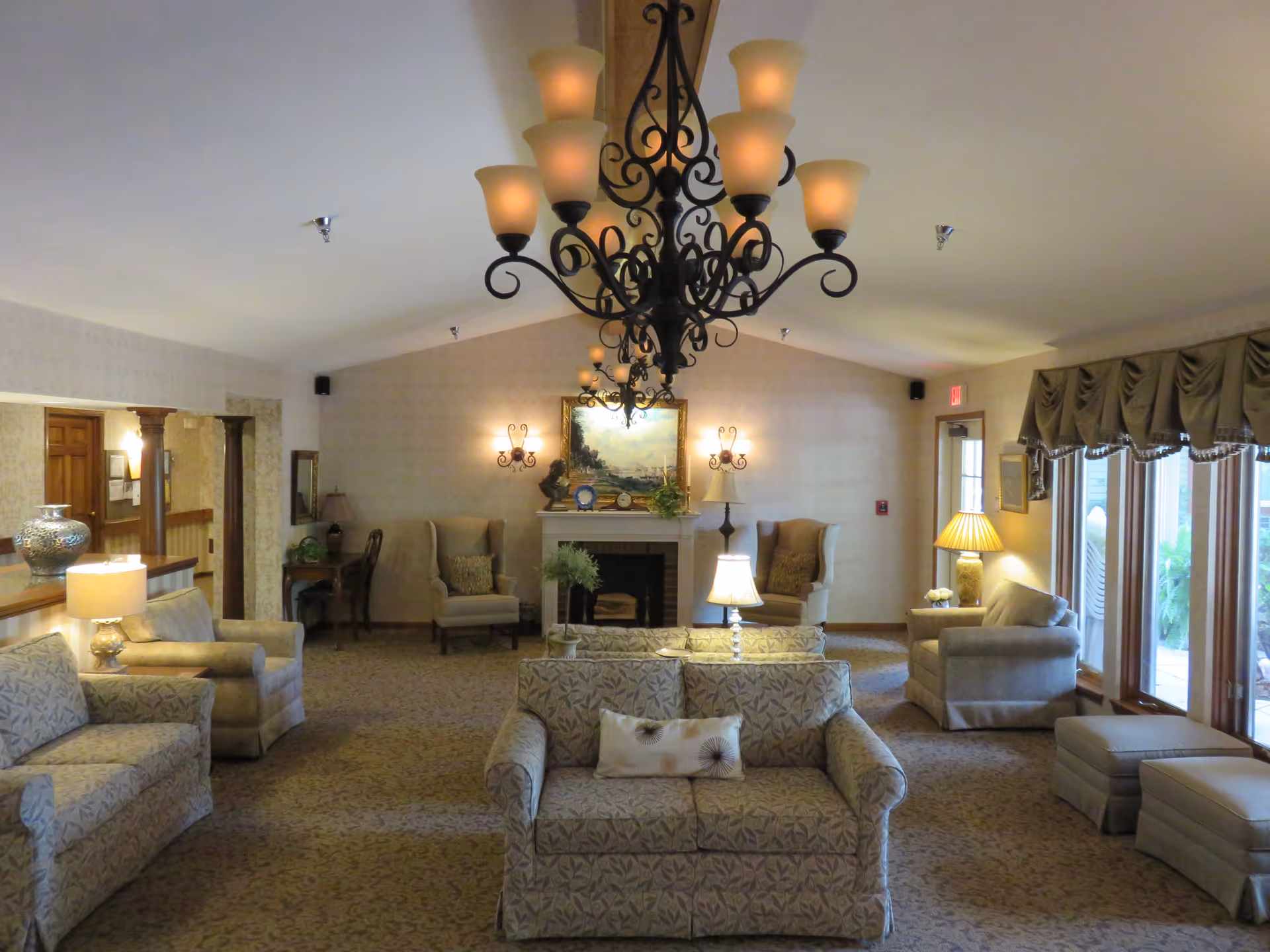 A cozy senior living community lounge with patterned sofas and armchairs arranged around a central fireplace. The room features warm lighting from table lamps and wall sconces, a large decorative chandelier hanging from the ceiling, and large windows with draped curtains letting in natural light.