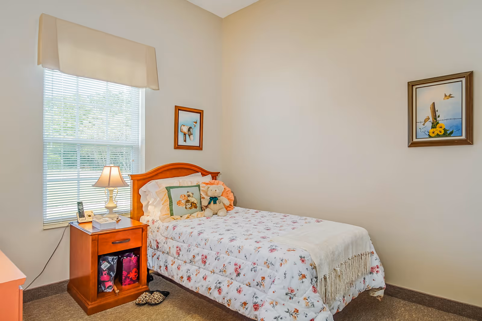 A small bedroom with a single bed covered in a floral bedspread and a white throw blanket. There is a teddy bear and a decorative pillow on the bed. Next to the bed is a wooden nightstand with a lamp, a phone, and a tissue box. A window with blinds and a beige valance is behind the nightstand. Two framed pictures hang on the walls, one above the bed and one on the adjacent wall. The floor is carpeted and there is a pair of slippers on the floor near the nightstand.