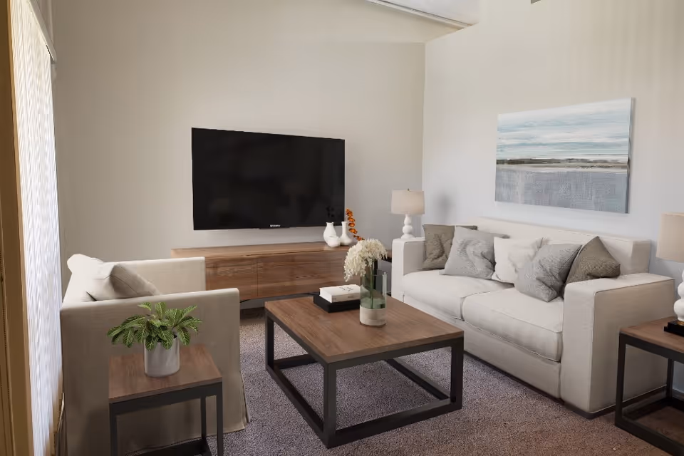 A modern living room with a beige sofa and armchair, a wooden coffee table with a vase of flowers and books, a wooden TV stand with decorative items, a large flat-screen TV mounted on the wall, two table lamps, and a landscape painting on the wall.