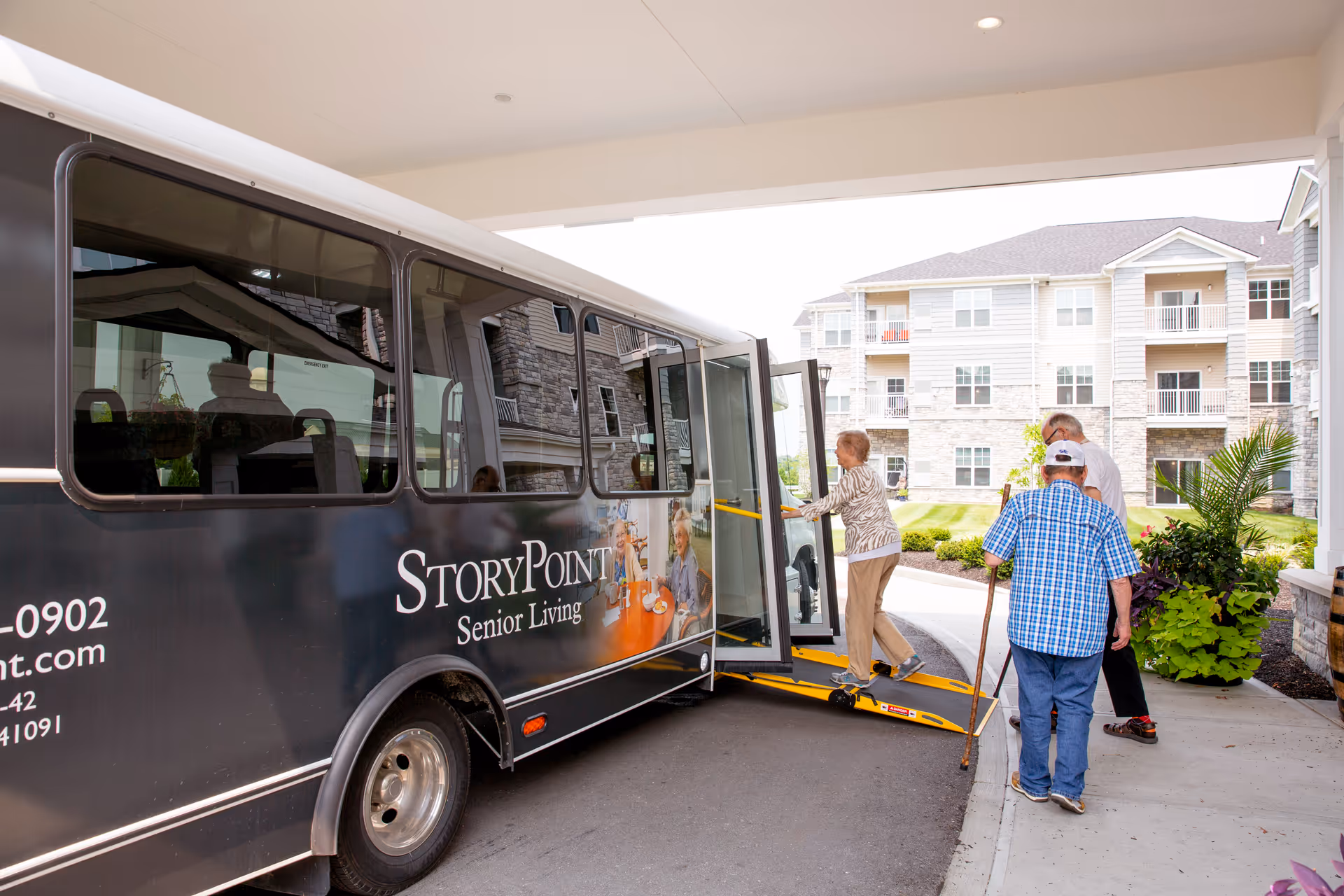 Three elderly individuals boarding a StoryPoint Senior Living shuttle bus using a wheelchair ramp outside a senior living facility with multi-story residential buildings and landscaped greenery in the background.