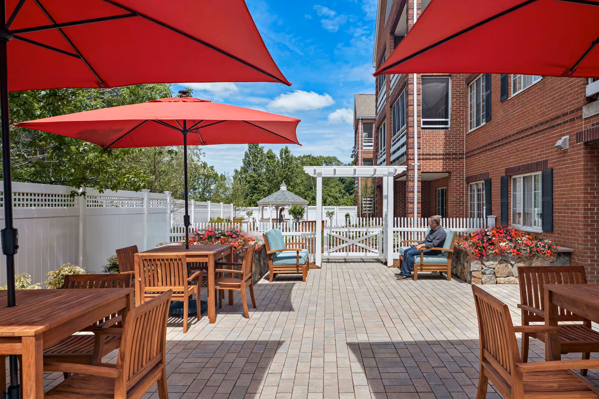 Outdoor patio courtyard with wooden tables and red umbrellas next to a brick building and white picket fence.