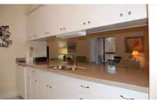 Interior view of a kitchen area with white cabinets and drawers, a countertop with a sink, and a faucet. In the background, there is a dining area with a table, chairs, a lamp, and framed artwork on the wall.