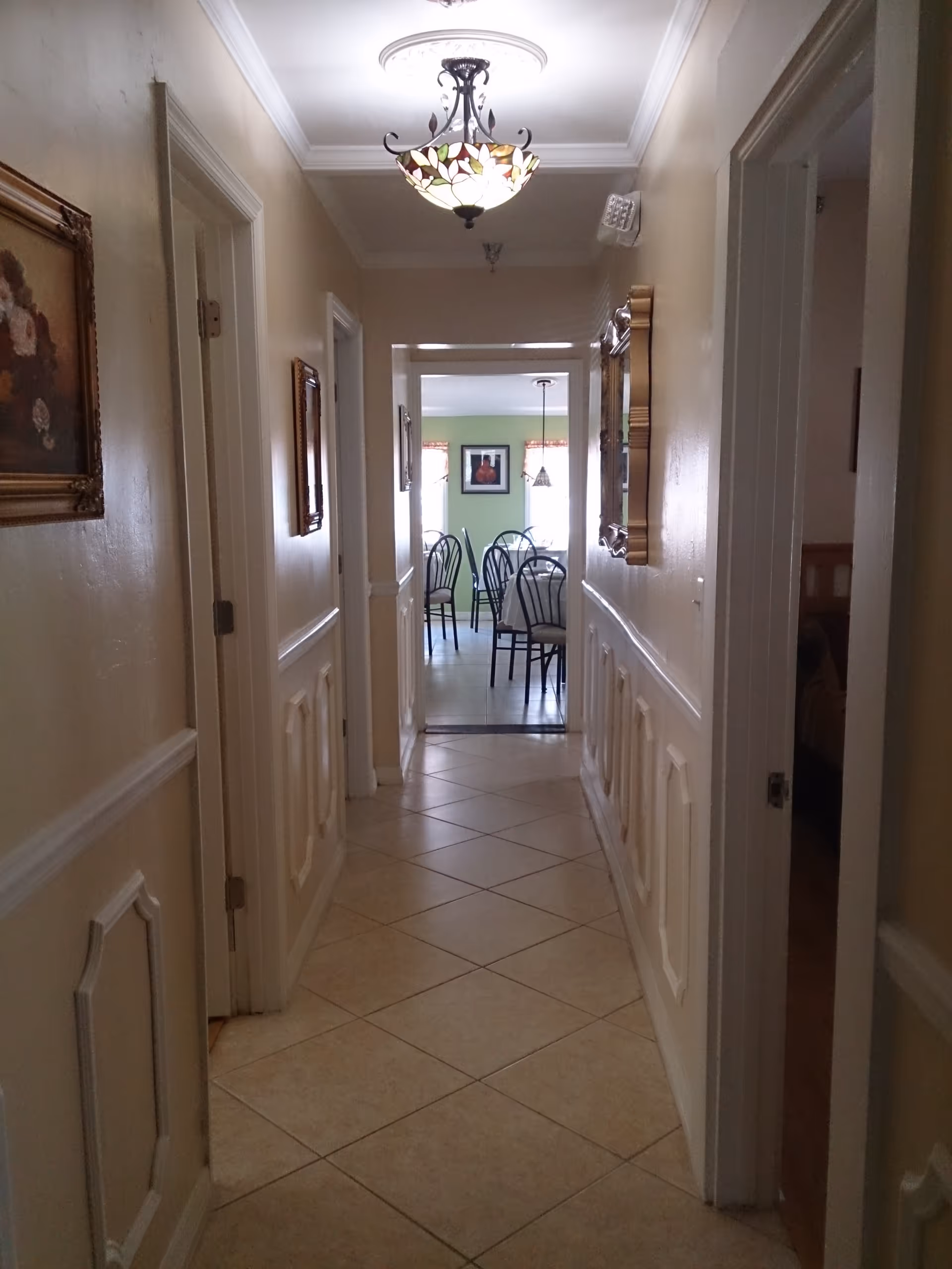 A narrow hallway with beige walls and tiled floor leading to a dining area with a table and chairs. The hallway has decorative wall panels, framed pictures on the walls, and a stained glass ceiling light fixture. Doors to other rooms are visible on both sides of the hallway.