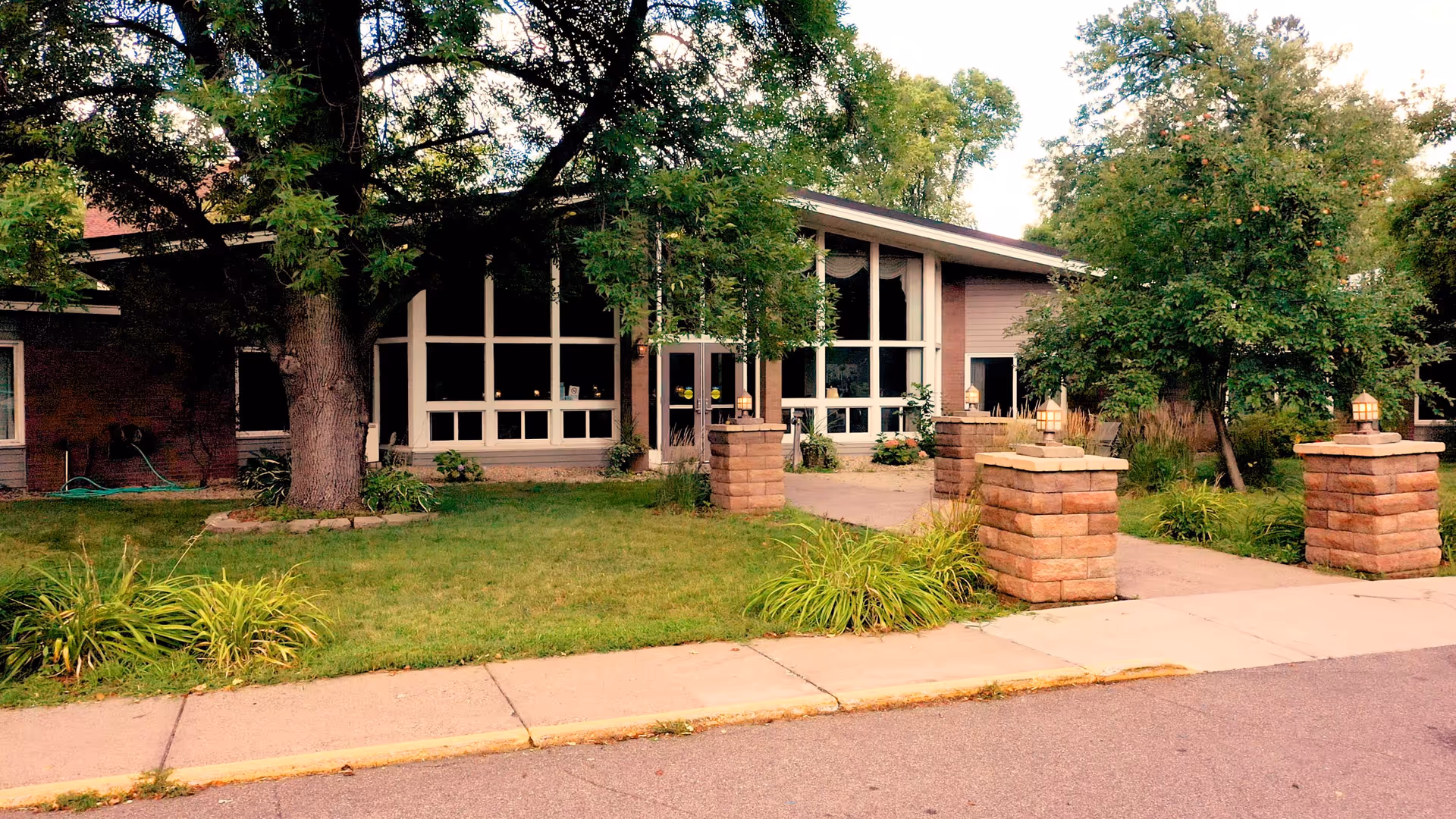 Front entrance of a single-story senior living facility with large windows, lawn, trees, and stone pillars flanking the walkway.