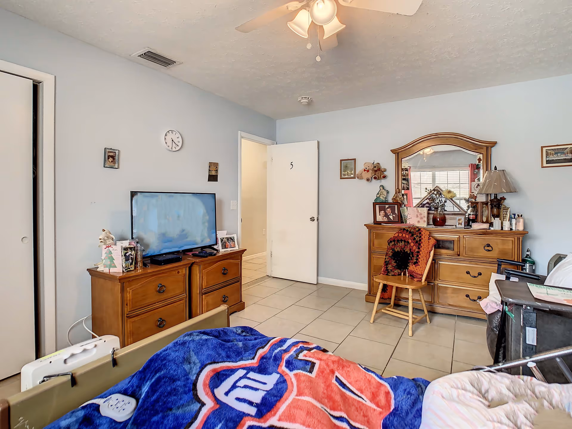 Interior view of a bedroom in a senior living facility with light blue walls, a ceiling fan, and tiled floor. The room contains a wooden dresser with a flat-screen TV on top, a wooden dresser with a mirror and various personal items, a wooden chair with a crocheted blanket draped over it, and a bed partially visible with a blue and red blanket. The door to the room is open, showing a hallway outside.