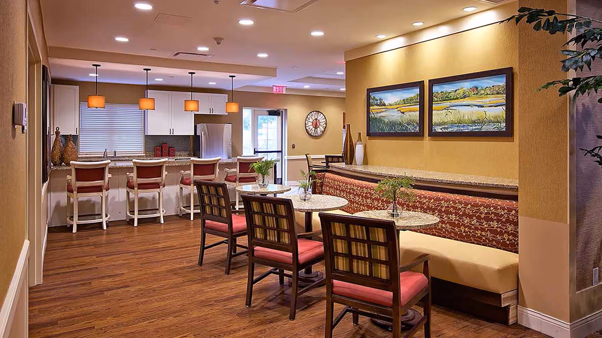 A cozy dining area in a senior living facility featuring round tables with chairs and a cushioned bench along the wall. The space has warm yellow walls, wooden flooring, and decorative paintings. In the background, there is a kitchen area with a counter, bar stools, pendant lights, and stainless steel appliances.
