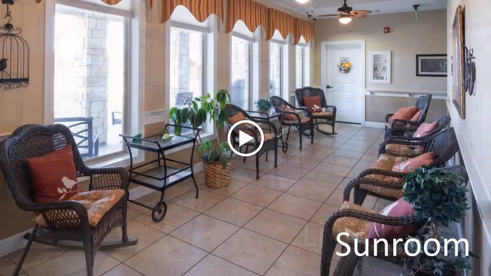 A sunroom with large windows along one wall letting in natural light. The room is furnished with multiple wicker chairs with cushions arranged along both sides. There are potted plants and a small rolling cart in the room. The floor is tiled, and the walls are light-colored with some decorative elements. A ceiling fan is visible, and a door is at the far end of the room.