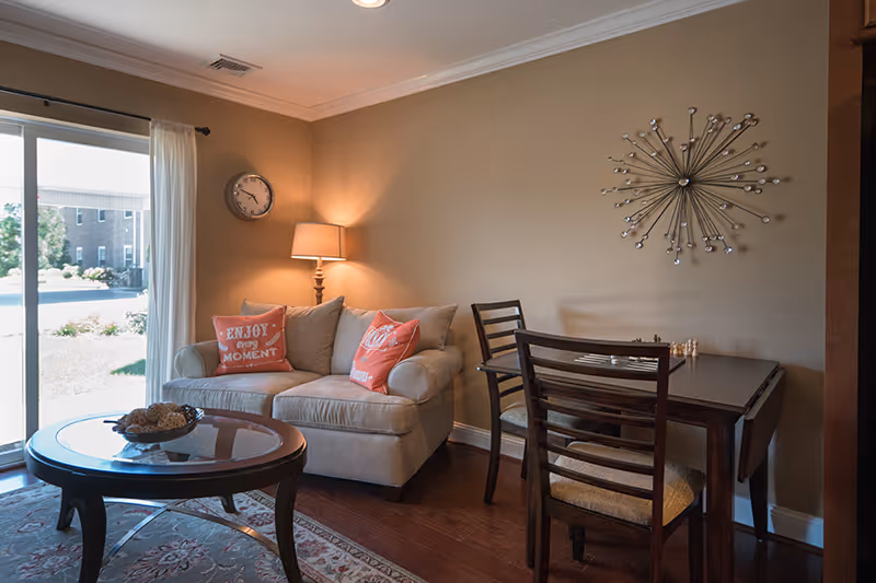 Cozy living room with a loveseat and decorative pillows, floor lamp, round coffee table, and a small dining table by a sliding glass door.