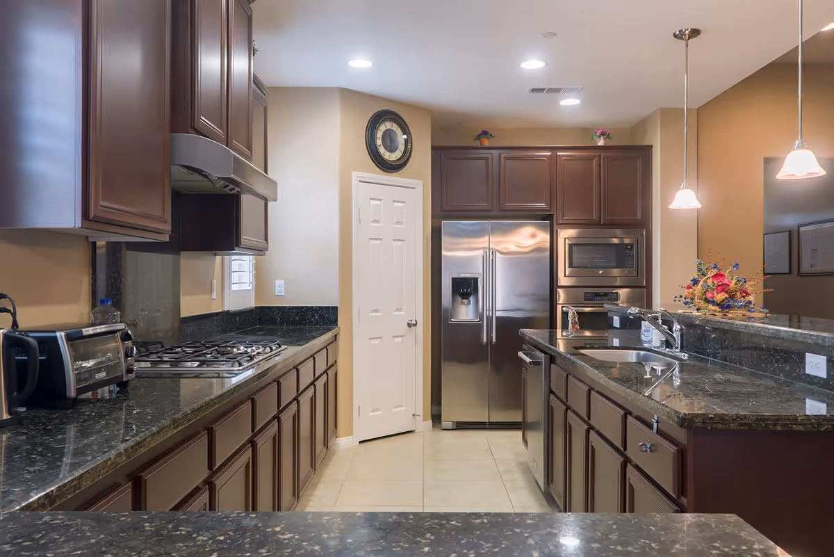 A modern kitchen with dark wood cabinets, black granite countertops, stainless steel appliances including a refrigerator, microwave, and dishwasher. The kitchen features a gas stove, a toaster oven, a sink with a faucet, and pendant lights hanging from the ceiling. There is a clock on the wall above a white door, and a floral arrangement on the counter.