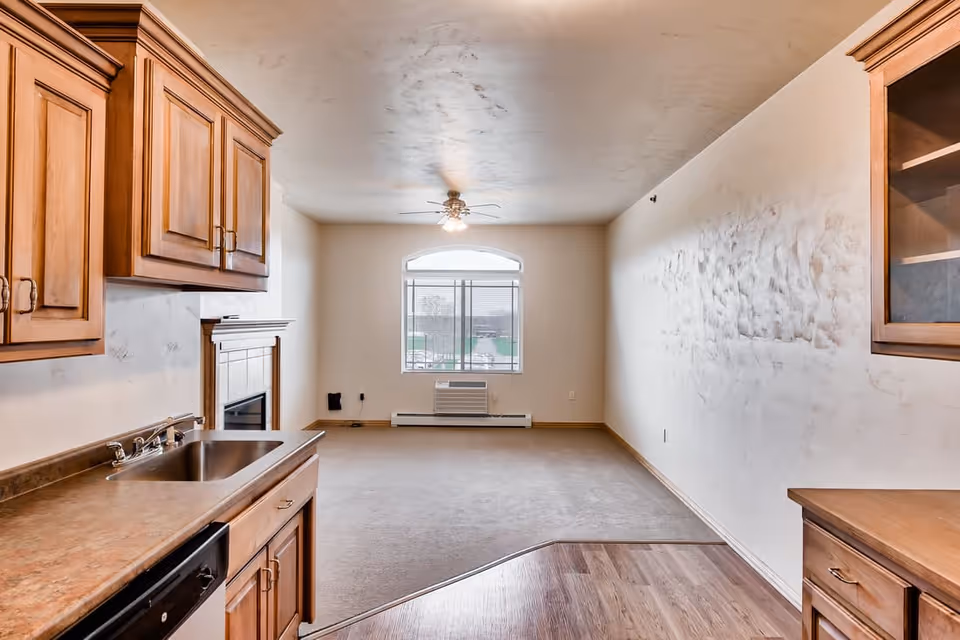 Interior view of a senior living apartment featuring a kitchen area with wooden cabinets and a sink on the left, transitioning into a carpeted living room with a ceiling fan and a large window with blinds. There is a fireplace on the left wall and an air conditioning unit below the window.