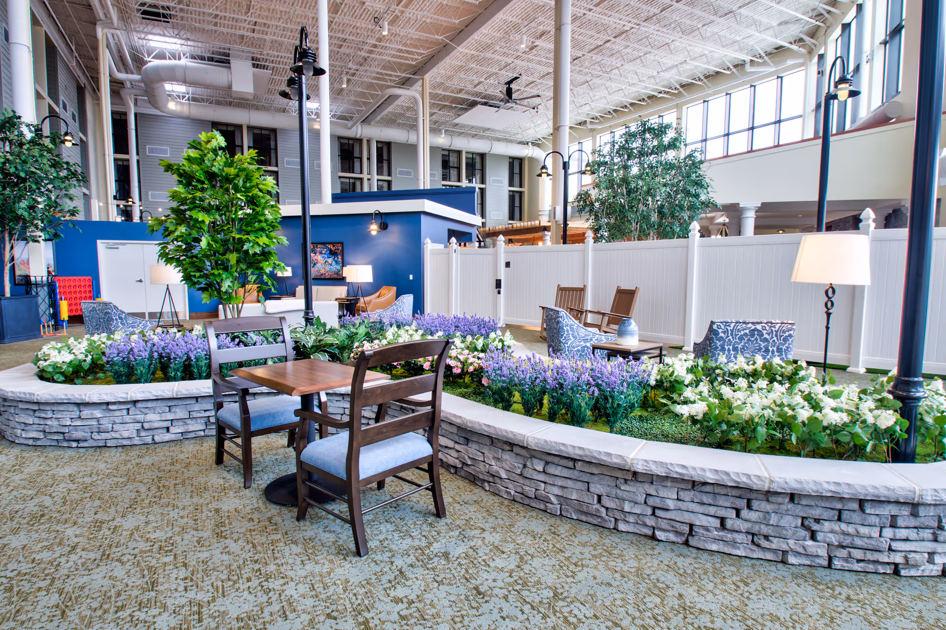Indoor seating area at Prosper at Wickliffe featuring a raised stone planter filled with purple and white flowers, surrounded by wooden chairs and small tables. The space has high ceilings with exposed beams, large windows letting in natural light, tall indoor trees, and modern floor lamps providing additional lighting.