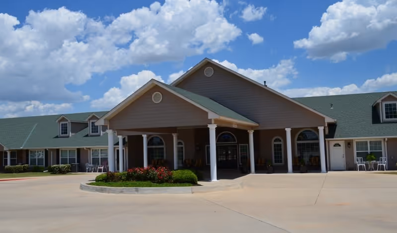 Front exterior view of Hefner Mansions Senior Independent Living building with a covered entrance supported by white columns, green roof, and a clear blue sky with scattered clouds.