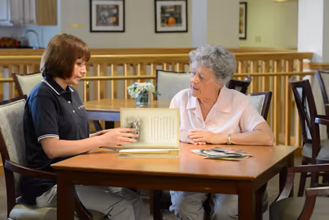 A staff member and an elderly resident sit at a table in a senior living facility, looking through a photo album in a dining/common area.